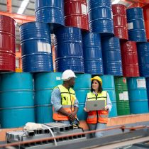 Multi-ethnic team of technicians wearing protective helmets studying operating manual while standing at CNC machine unit